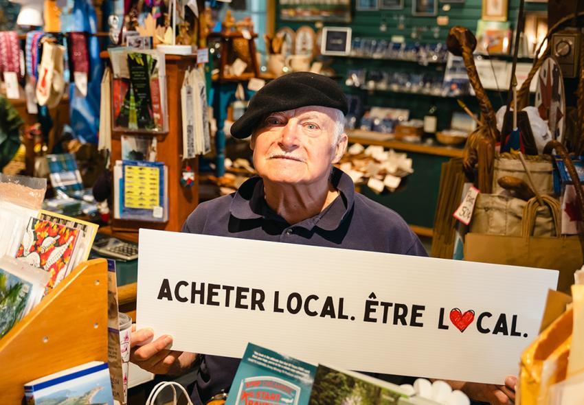 Michel Levasseur, owner of Carrefour Atlantic Emporium and the Puffin Gallery, holding a sign in French "Acheter Local. Ètre Local."