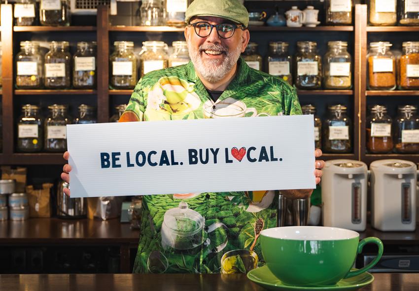 Phil Holmans, owner of World Tea House, holding a sign "Be Local. Buy Local."