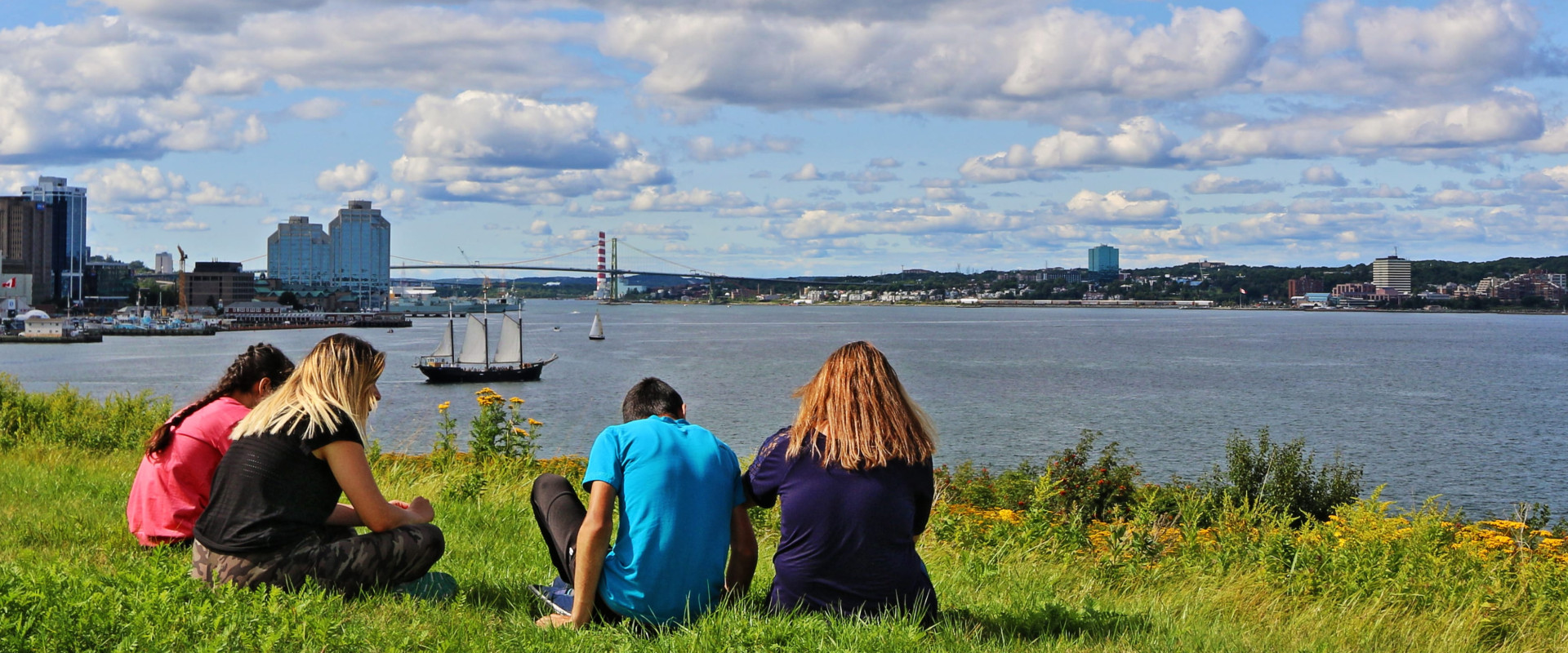 Three youth sitting on the grass on Georges Island looking towards the Halifax Waterfront.