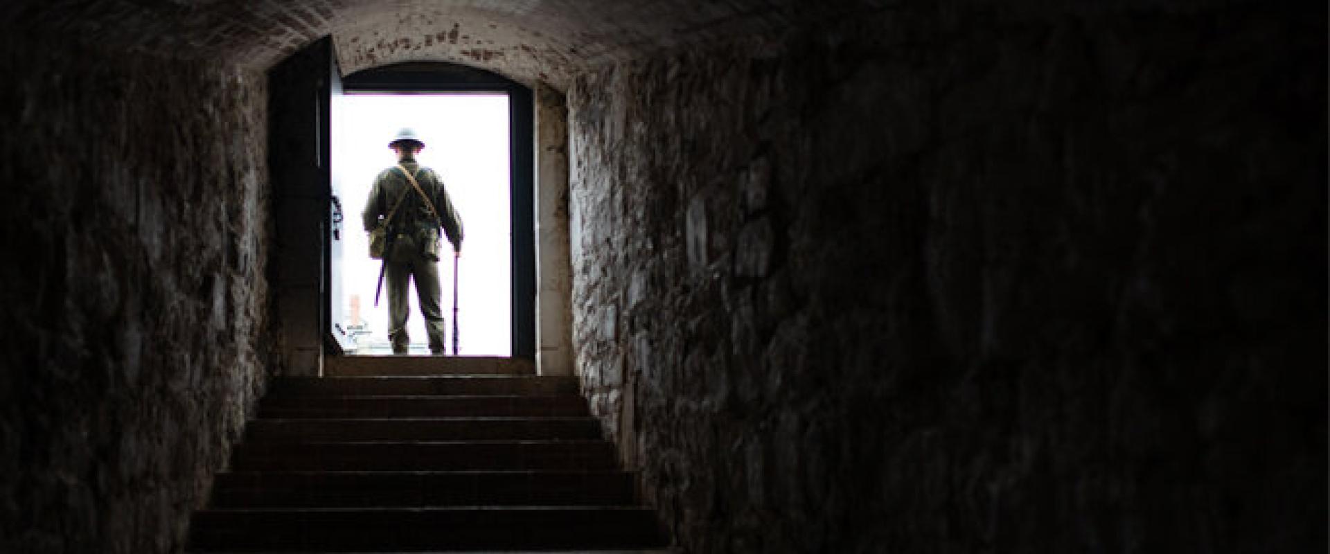 Photograph of a dark stairwell located within the Halifax Citadel National Historic Site fortress walls. At the end is a bright doorway where stands an interpreter in World War 2 uniform.