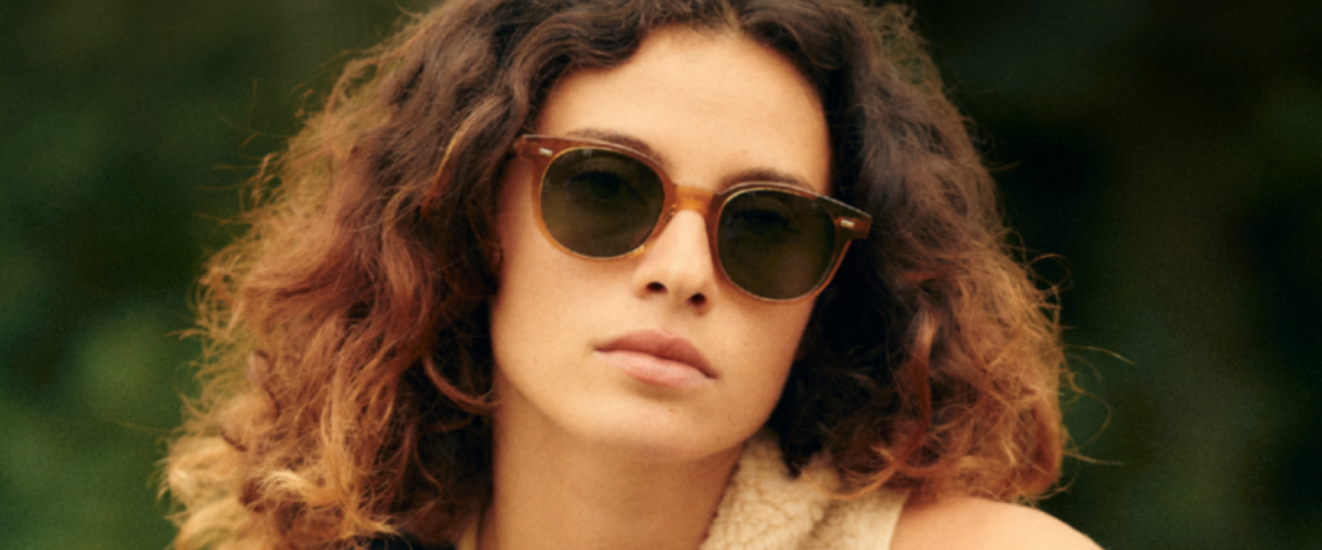 A woman with curly brown hair and a sherpa vest relaxes in an outdoor scene with amber colored sunglasses on.