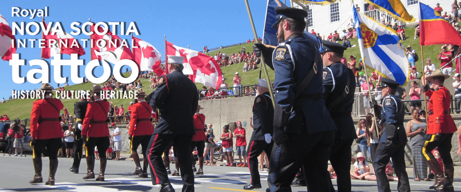 Royal Nova Scotia International Tattoo logo. RCMP members in red serge hold Canadian flags and Halifax Regional Police members in uniform hold flags while marching in a parade past the Town Clock on Citadel Hill. Crowds on the hill watch the parade go by on a sunny day.