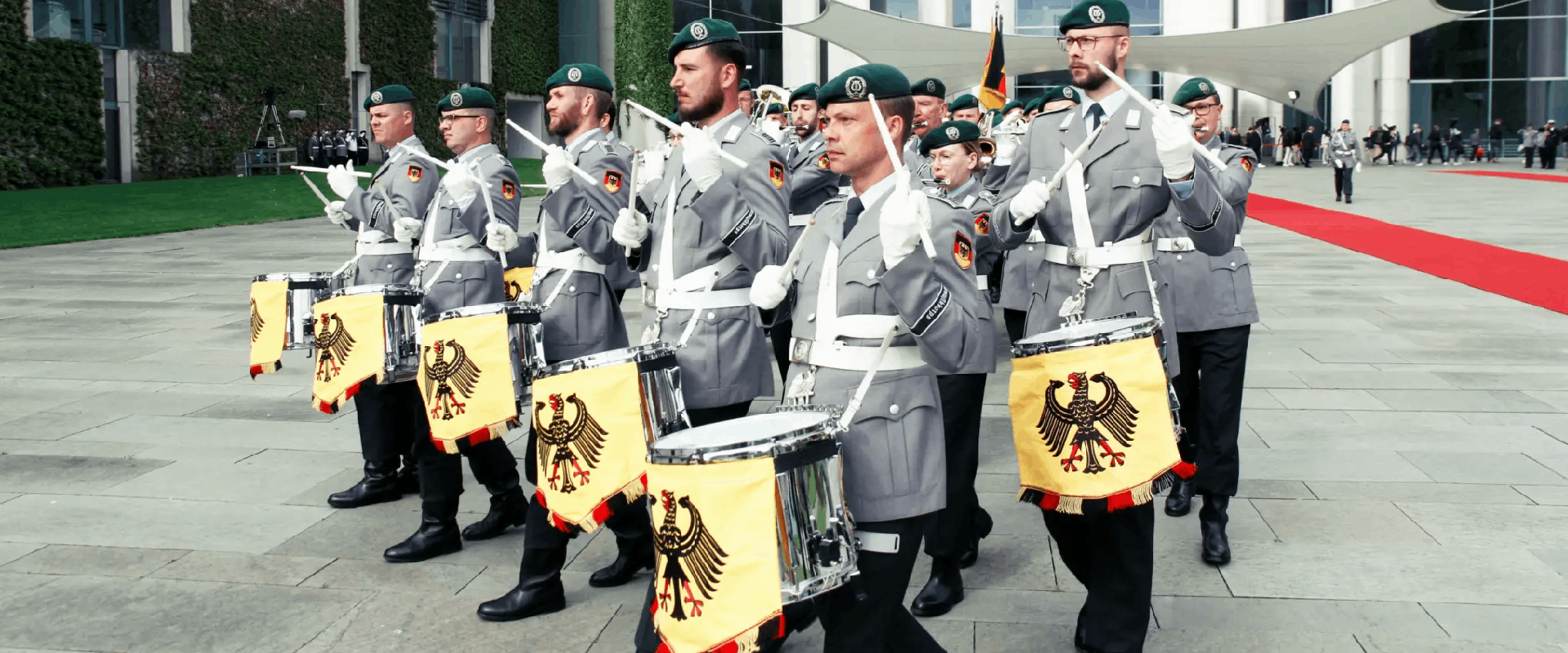 Drummers lead the Bundeswehr Ceremonial Band marching down a street. They wear light grey uniforms with dark green berets.