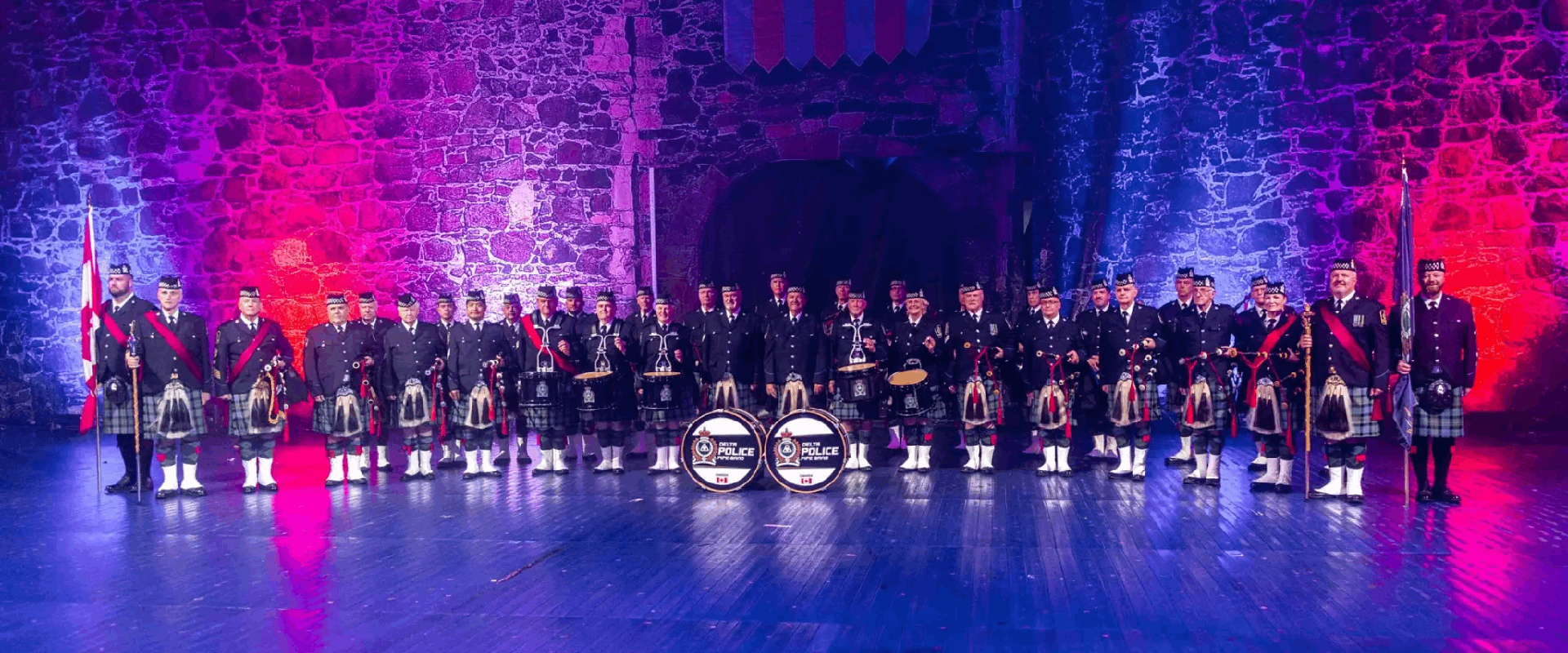 The Delta Police Pipe band stand in uniform in front of a castle wall with blue and red lighting.