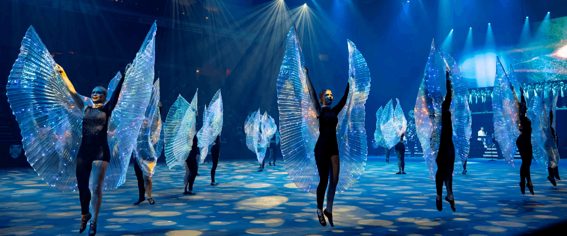 Highland dancers pose with arms high in black bodysuits with iridescent light up wings, in formation on an arena floor with blue lighting.