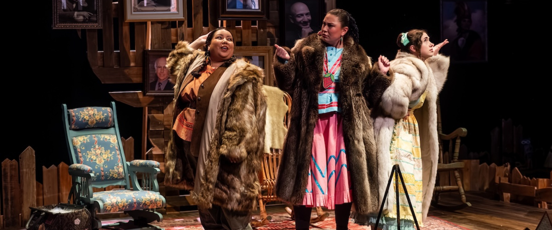 Three women - the titular "Women of the Fur Trade" - stand in glamorous poses on a stage set like a living room, complete with portraits, rocking chairs, and a rug. They wear long fur coats over their colourful outfits.