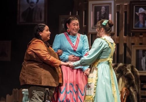 Three women face each other and hold hands, wearing colourful outfits. They are on a stage set like a living room, complete with portraits, rocking chairs, and a rug.