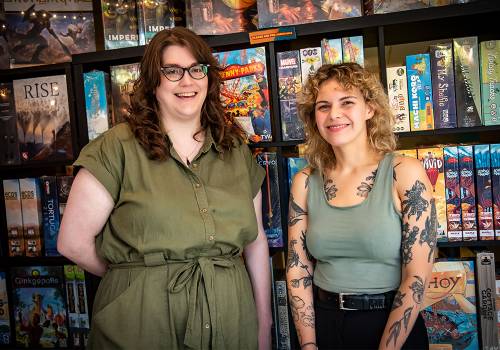 Kat and Brittney, managers of the Board Room Game Cafe posing in front of a wall of board games