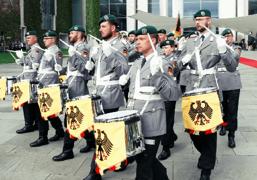 Drummers lead the Bundeswehr Ceremonial Band marching down a street. They wear light grey uniforms with dark green berets.
