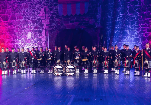 The Delta Police Pipe band stand in uniform in front of a castle wall with blue and red lighting.