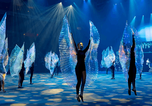 Highland dancers pose with arms high in black bodysuits with iridescent light up wings, in formation on an arena floor with blue lighting.