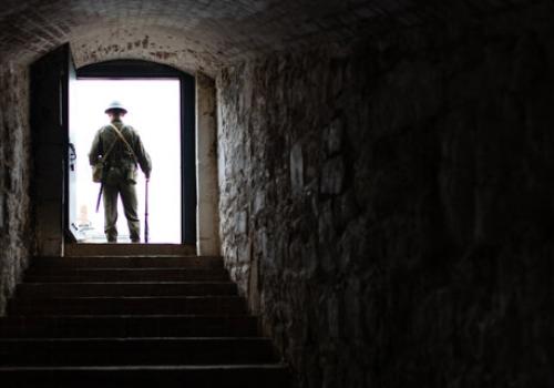 Photograph of a dark stairwell located within the Halifax Citadel National Historic Site fortress walls. At the end is a bright doorway where stands an interpreter in World War 2 uniform.