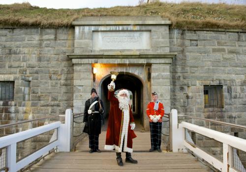 Father Christmas ringing a bell at the entrance of the Halifax Citadel fort with two costumed interpreters standing behind on either side.